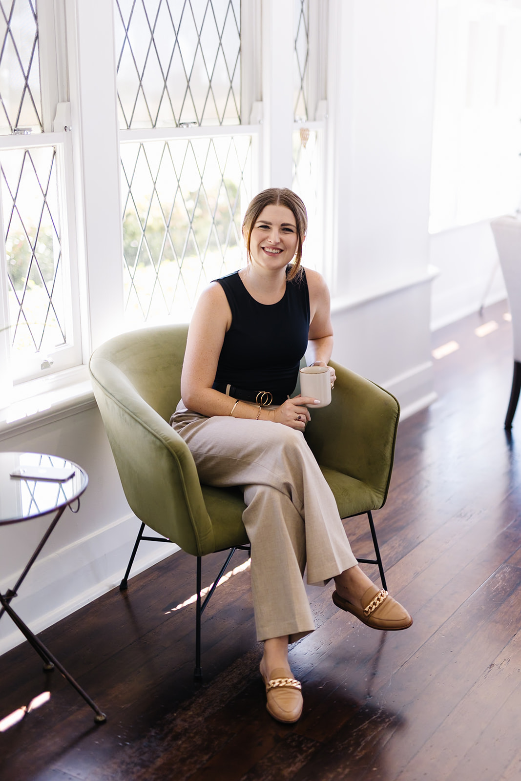 Ashleigh sitting on green velvet armchair holding coffee cup and smiling at camera.