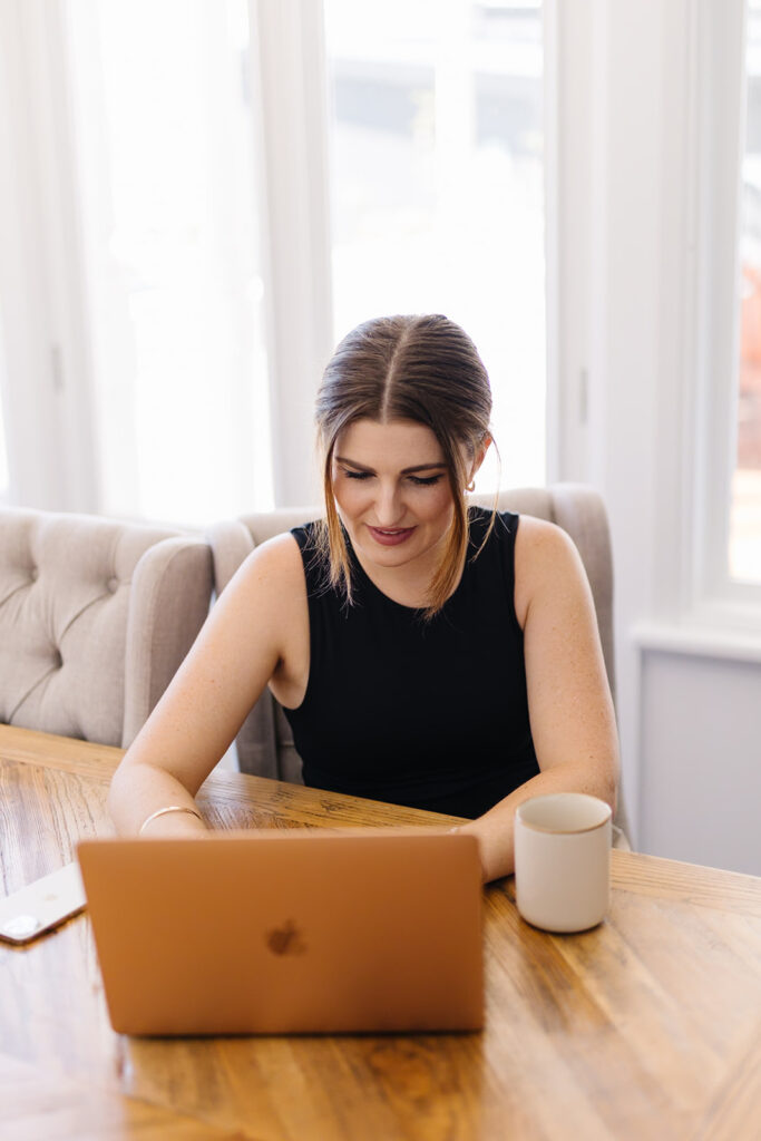 Ashleigh sitting at Apple Laptop smiling while typing with a cup of tea